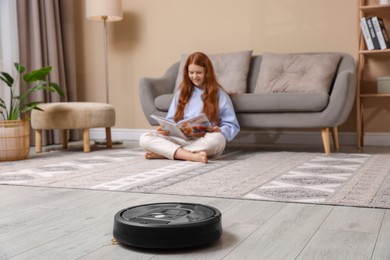 Photo of Teenage girl reading magazine while robotic vacuum cleaner vacuuming rug at home, selective focus