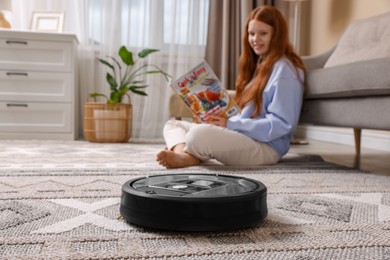 Photo of Teenage girl reading magazine while robotic vacuum cleaner vacuuming rug at home, selective focus