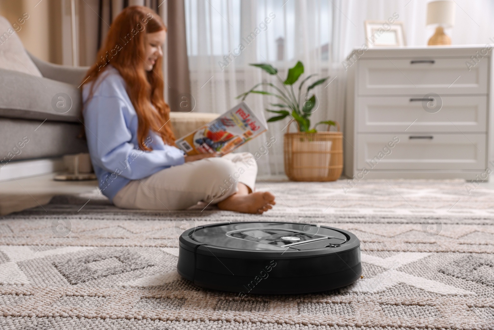 Photo of Teenage girl reading magazine while robotic vacuum cleaner vacuuming rug at home, selective focus