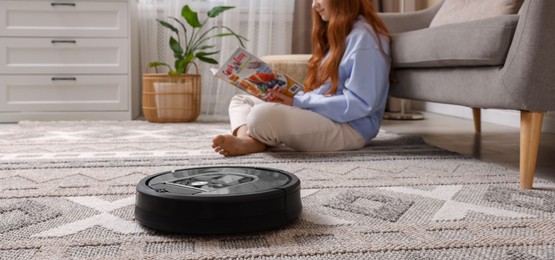 Photo of Teenage girl reading magazine while robotic vacuum cleaner vacuuming rug at home, selective focus