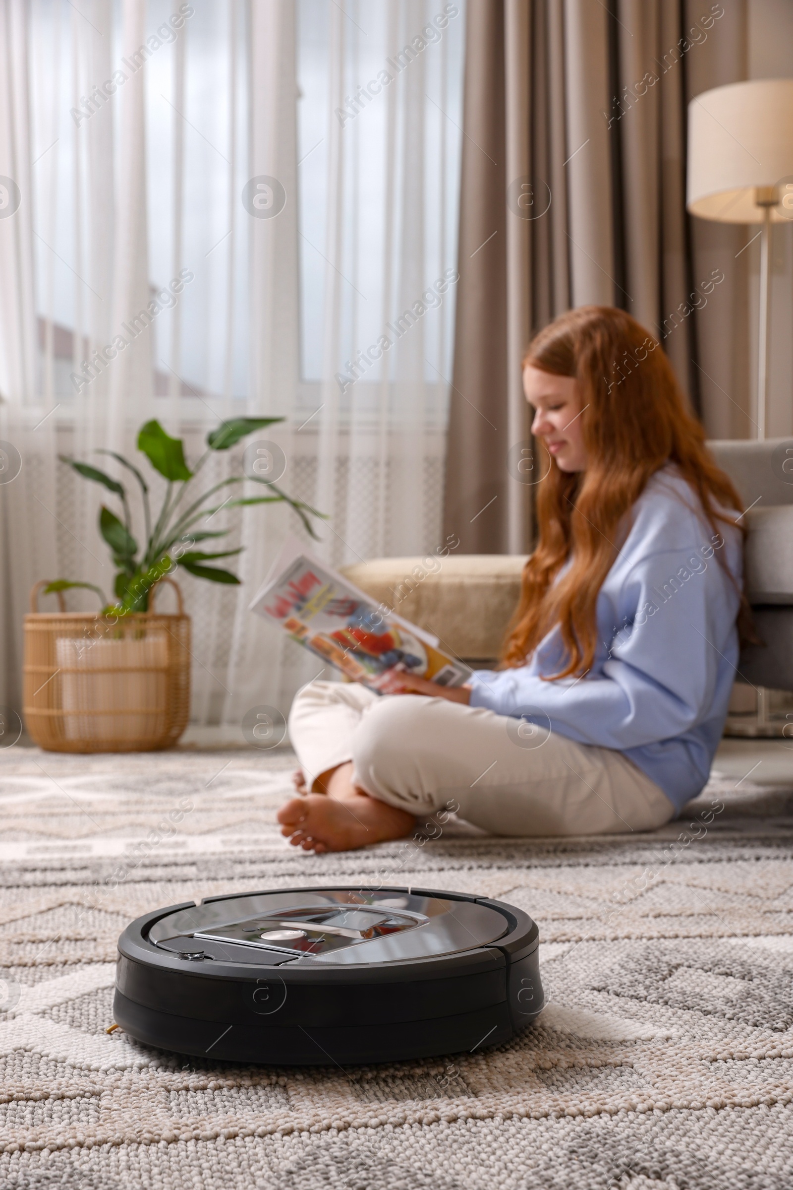 Photo of Teenage girl reading magazine while robotic vacuum cleaner vacuuming rug at home, selective focus