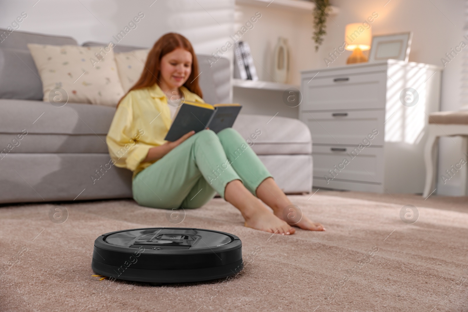 Photo of Teenage girl reading book while robotic vacuum cleaner vacuuming rug at home, selective focus