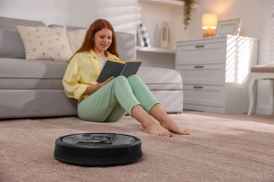 Photo of Teenage girl reading book while robotic vacuum cleaner vacuuming rug at home, selective focus