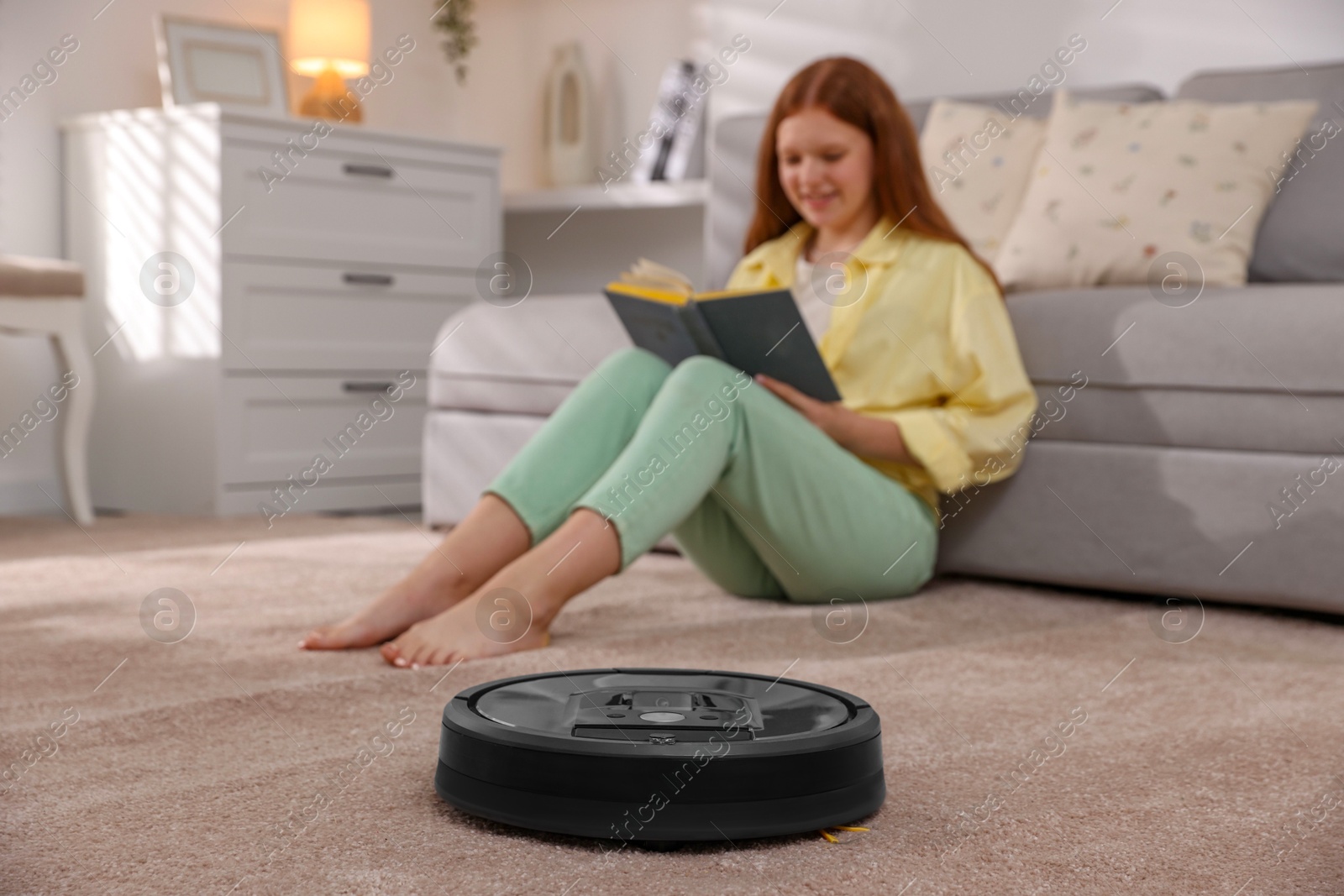 Photo of Teenage girl reading book while robotic vacuum cleaner vacuuming rug at home, selective focus