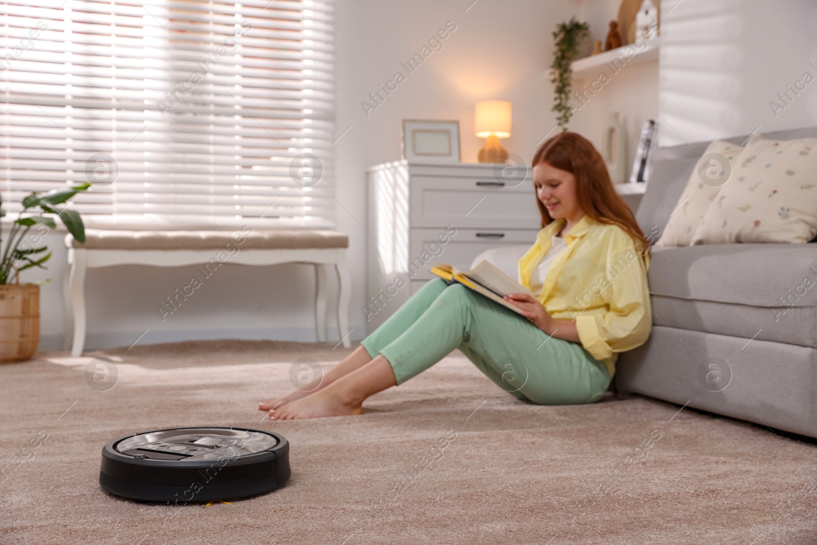 Photo of Teenage girl reading book while robotic vacuum cleaner vacuuming rug at home, selective focus