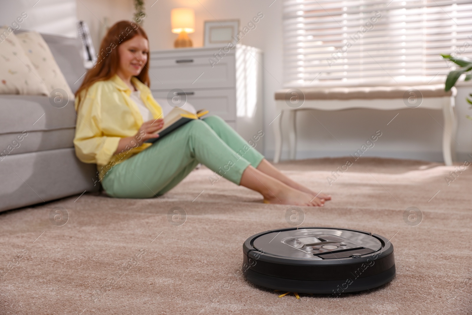 Photo of Teenage girl reading book while robotic vacuum cleaner vacuuming rug at home, selective focus