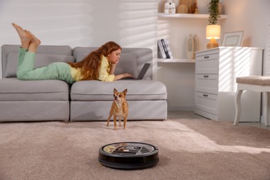 Photo of Teenage girl with laptop and her cute Chihuahua dog at home, focus on robotic vacuum cleaner