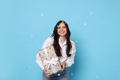 Photo of Happy woman with gift box under falling confetti on light blue background. Surprise party