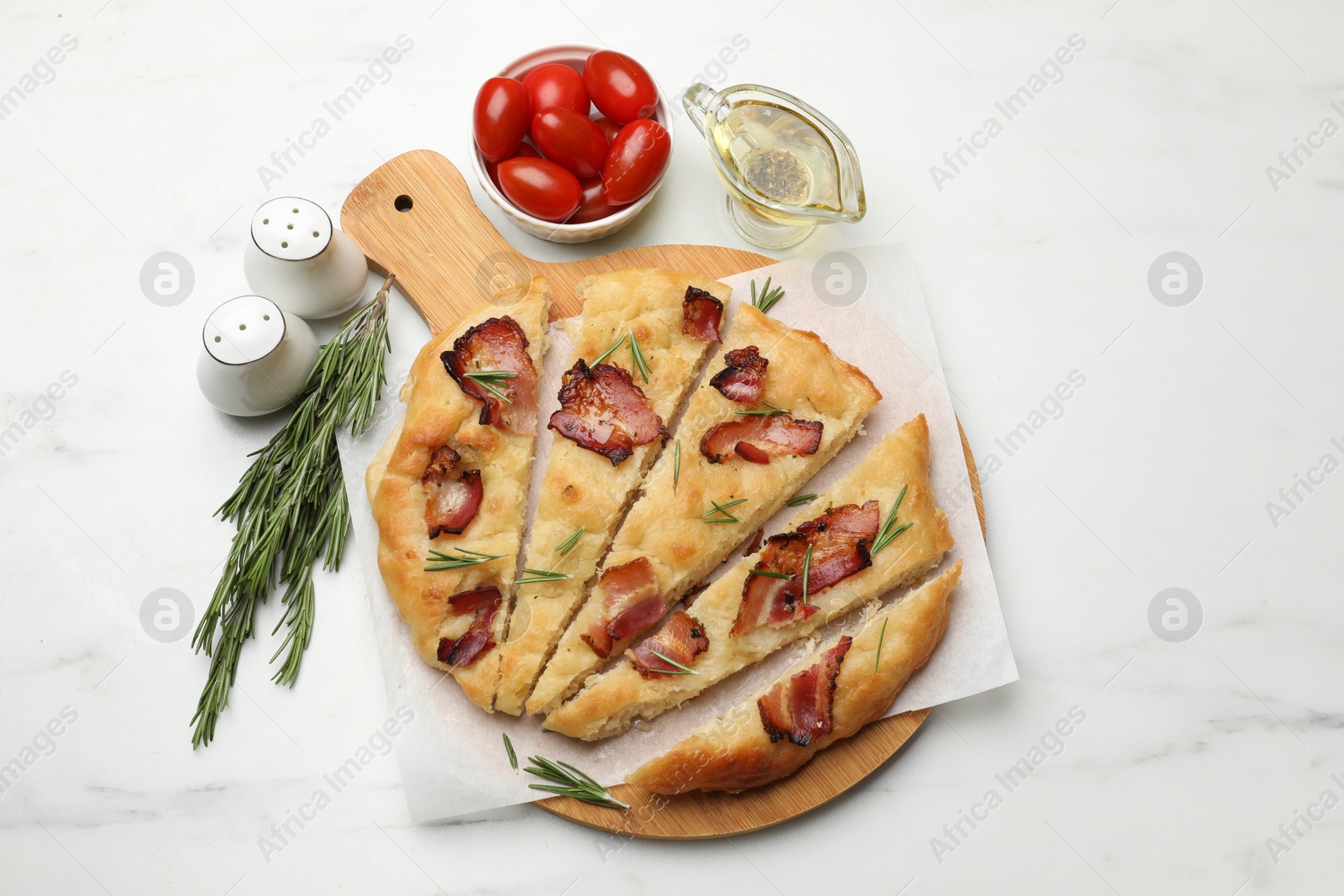 Photo of Slices of delicious focaccia bread with bacon, rosemary, oil and tomatoes on white marble table, flat lay