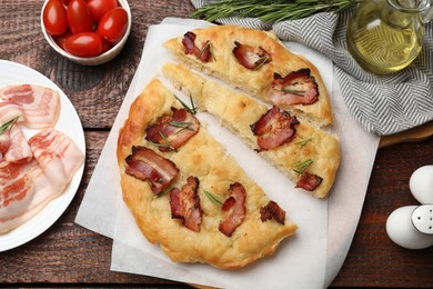 Photo of Slices of delicious focaccia bread with bacon, rosemary, oil and tomatoes on wooden table, flat lay