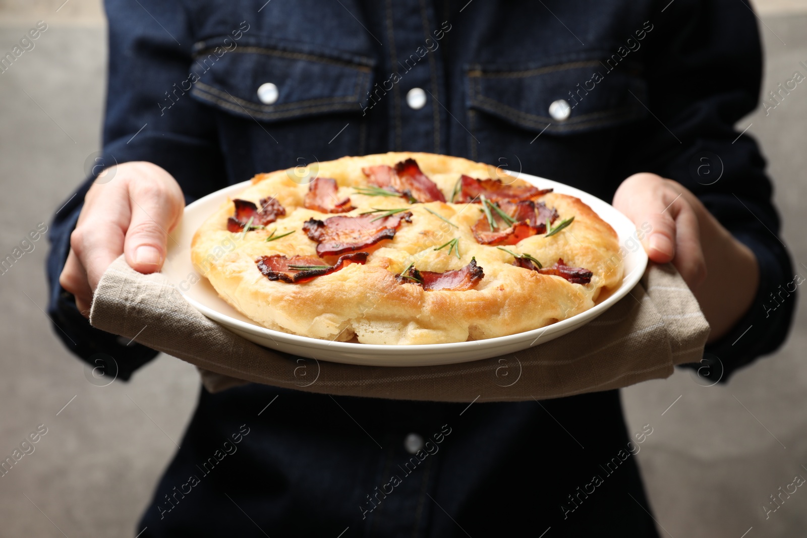 Photo of Woman holding delicious focaccia bread with bacon and rosemary on grey background, closeup