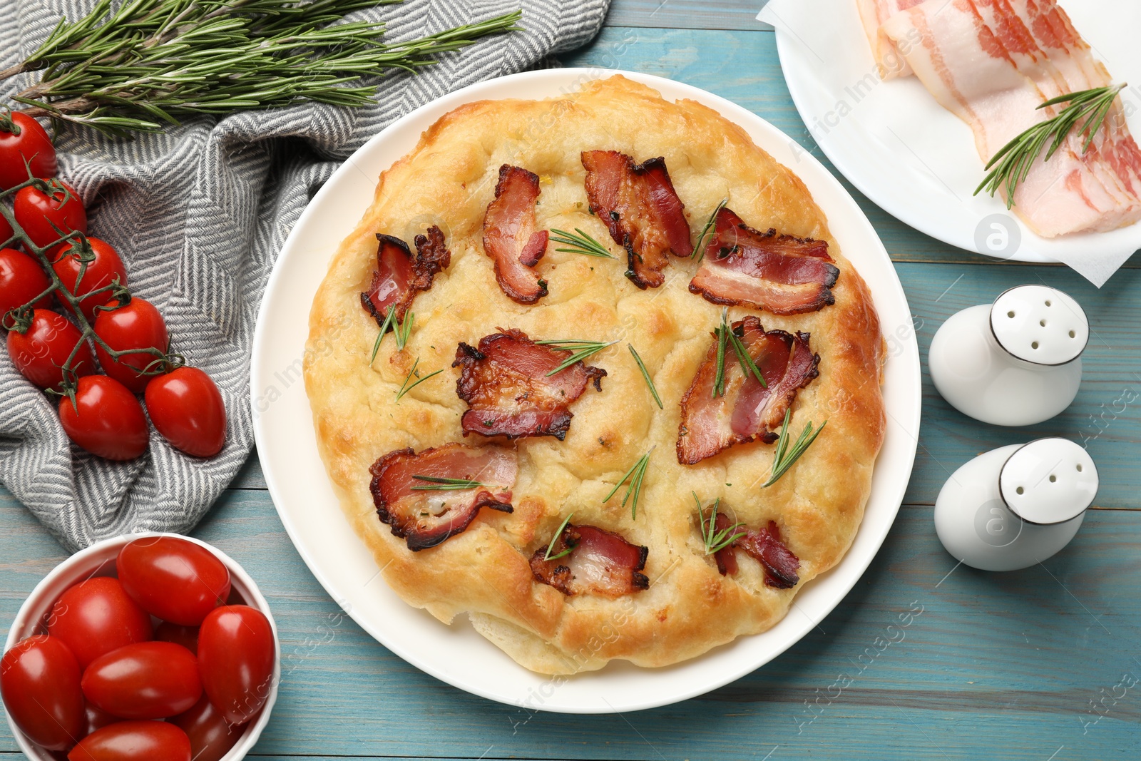 Photo of Delicious focaccia bread with bacon, rosemary and tomatoes on light blue wooden table, flat lay