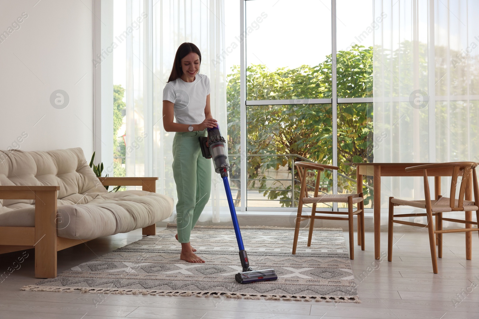 Photo of Smiling young woman cleaning rug with cordless vacuum cleaner at home