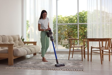 Smiling young woman cleaning rug with cordless vacuum cleaner at home