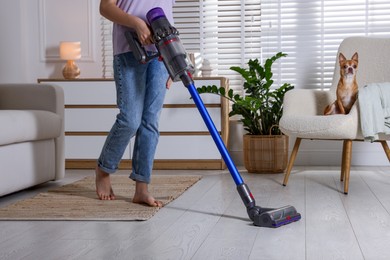 Photo of Teenage girl cleaning floor with cordless vacuum cleaner and her cute Chihuahua dog sitting in armchair at home, closeup