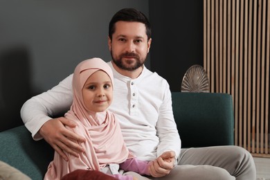 Photo of Muslim man and his daughter sitting on sofa at home