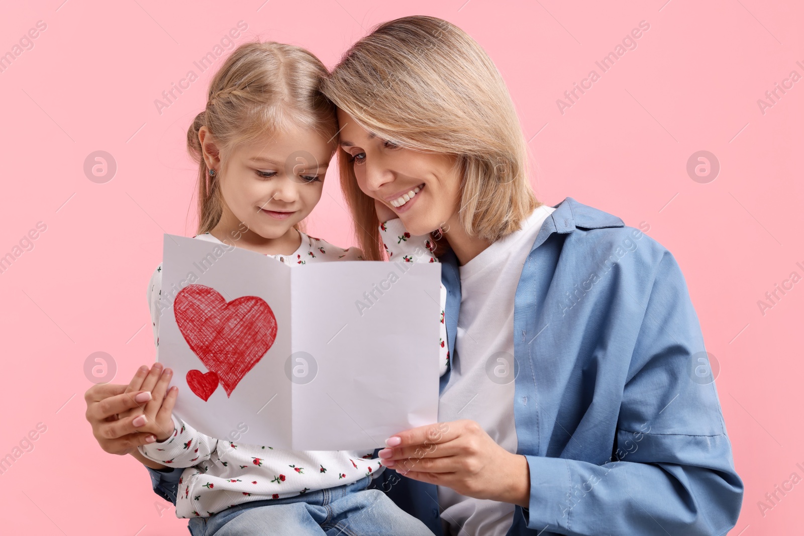 Photo of Happy woman with her daughter and greeting card on pink background. Mother's Day celebration