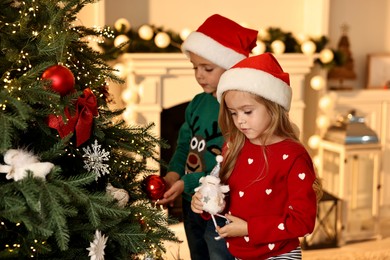 Photo of Little kids in Santa hats decorating Christmas tree at home