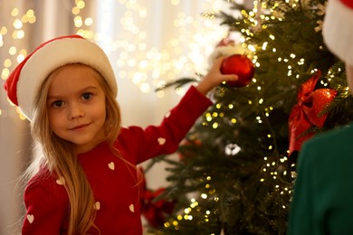 Photo of Little girl in Santa hat decorating Christmas tree at home