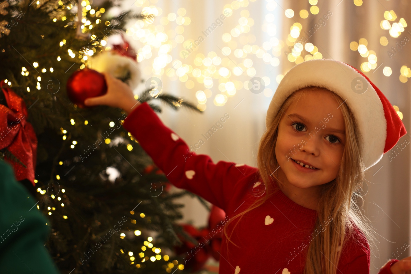 Photo of Little girl in Santa hat decorating Christmas tree at home