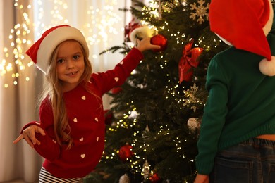 Photo of Little kids in Santa hats decorating Christmas tree at home