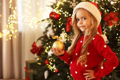 Photo of Little girl in Santa hat with Christmas ball at home, space for text