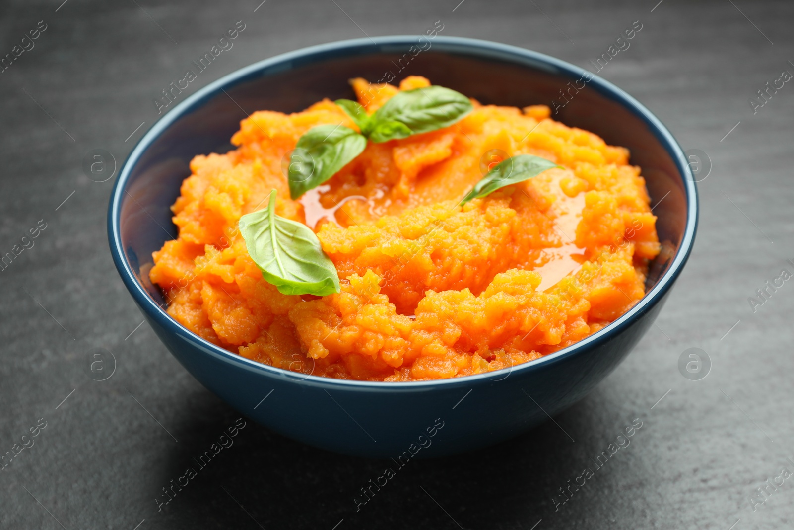 Photo of Tasty mashed sweet potato with basil in bowl on dark gray textured table, closeup