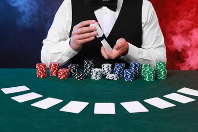 Photo of Professional croupier shuffling cards at gambling table against color background with smoke, closeup