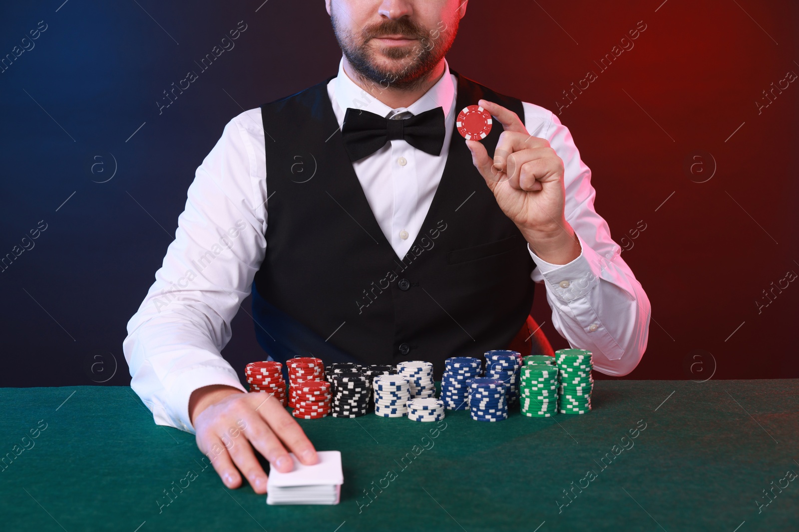 Photo of Professional croupier with playing cards and casino chips at gambling table against color background, closeup