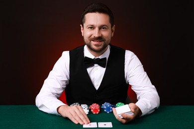 Photo of Professional croupier with playing cards at gambling table against dark red background