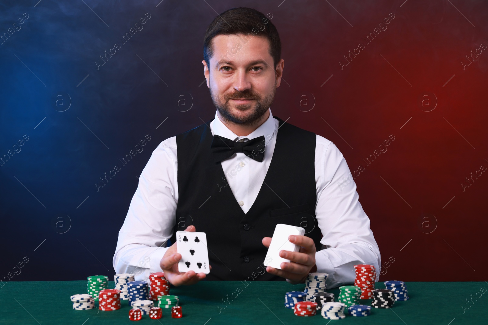 Photo of Professional croupier with playing cards at gambling table against color background with smoke