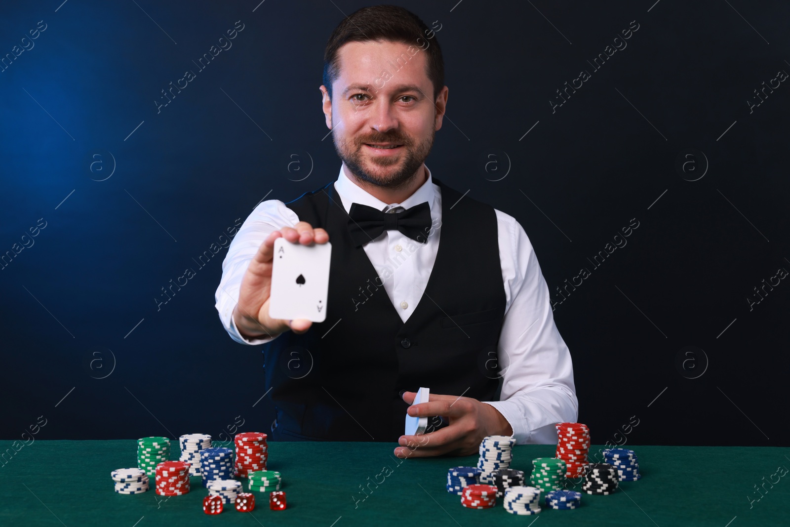 Photo of Professional croupier with playing cards at gambling table against dark blue background