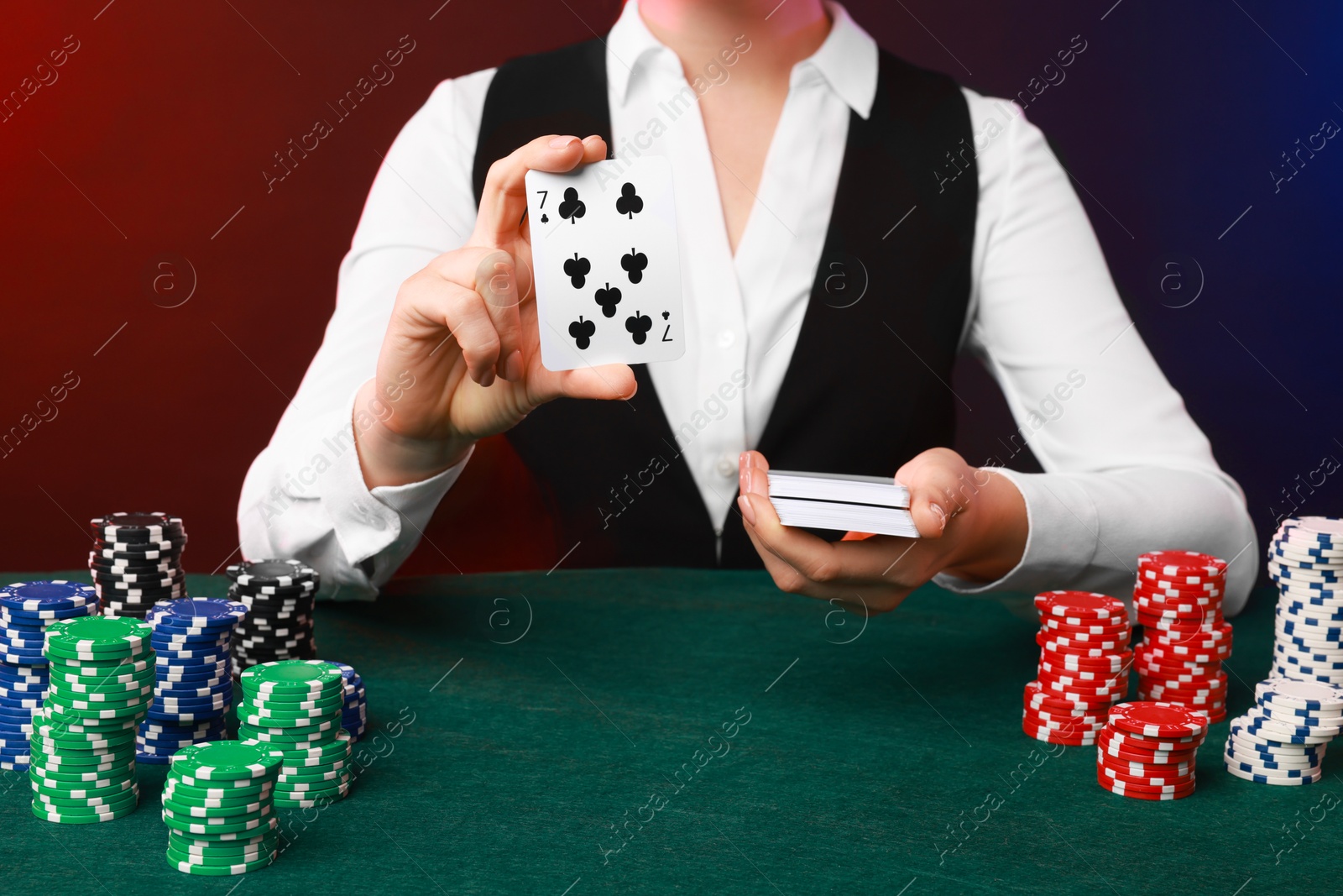 Photo of Professional croupier with casino chips and playing cards at gambling table on color background, closeup