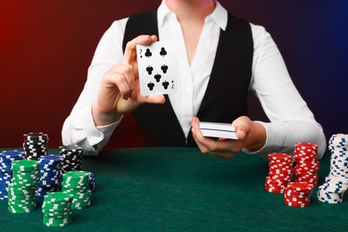 Photo of Professional croupier with casino chips and playing cards at gambling table on color background, closeup