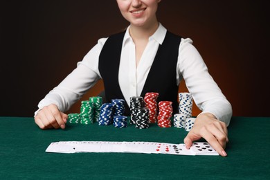 Photo of Professional croupier with casino chips and playing cards at gambling table on color background, closeup