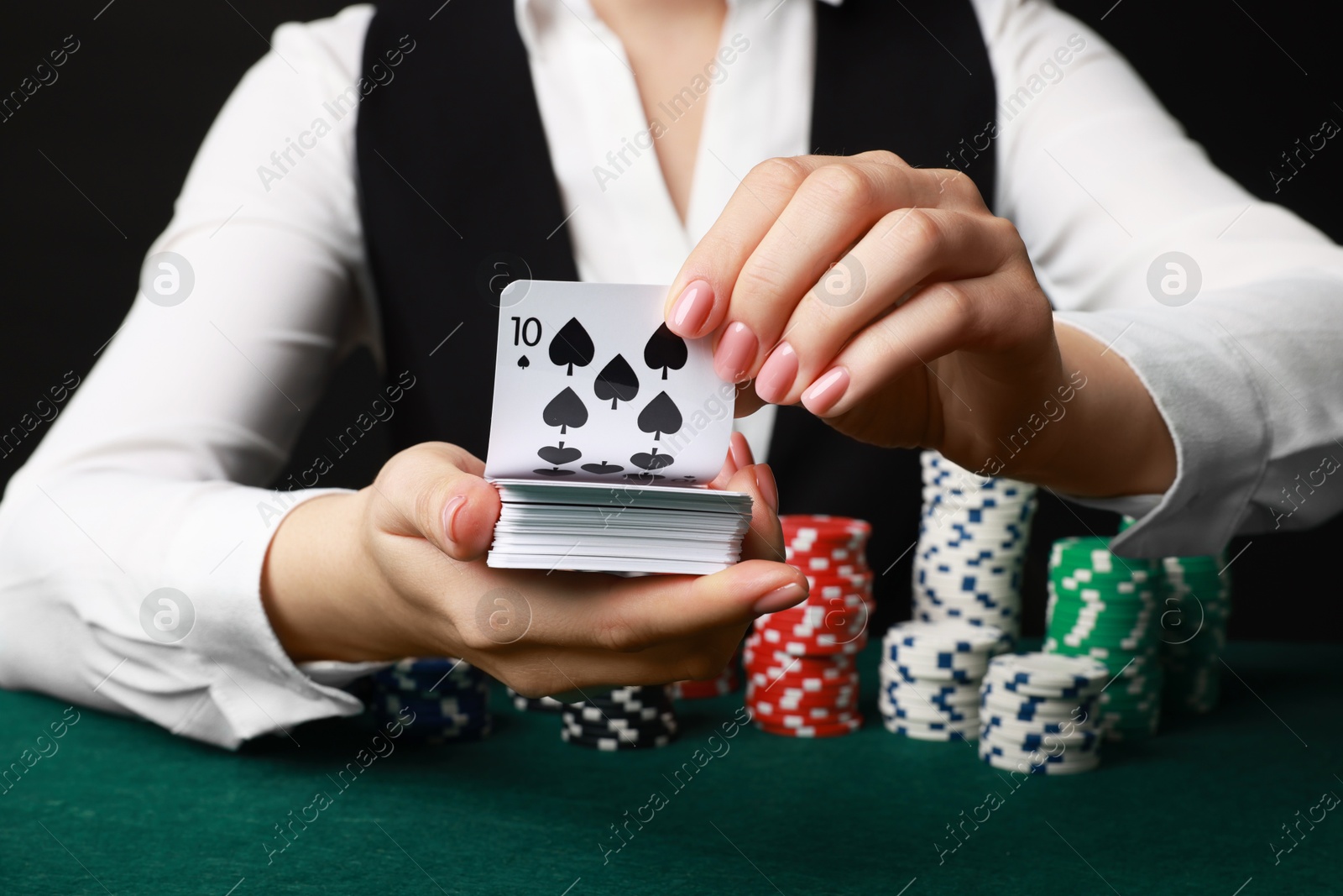 Photo of Professional croupier with chips shuffling playing cards at gambling table on black background, closeup