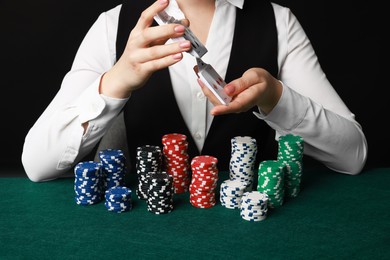 Photo of Professional croupier with chips shuffling playing cards at gambling table on black background, closeup