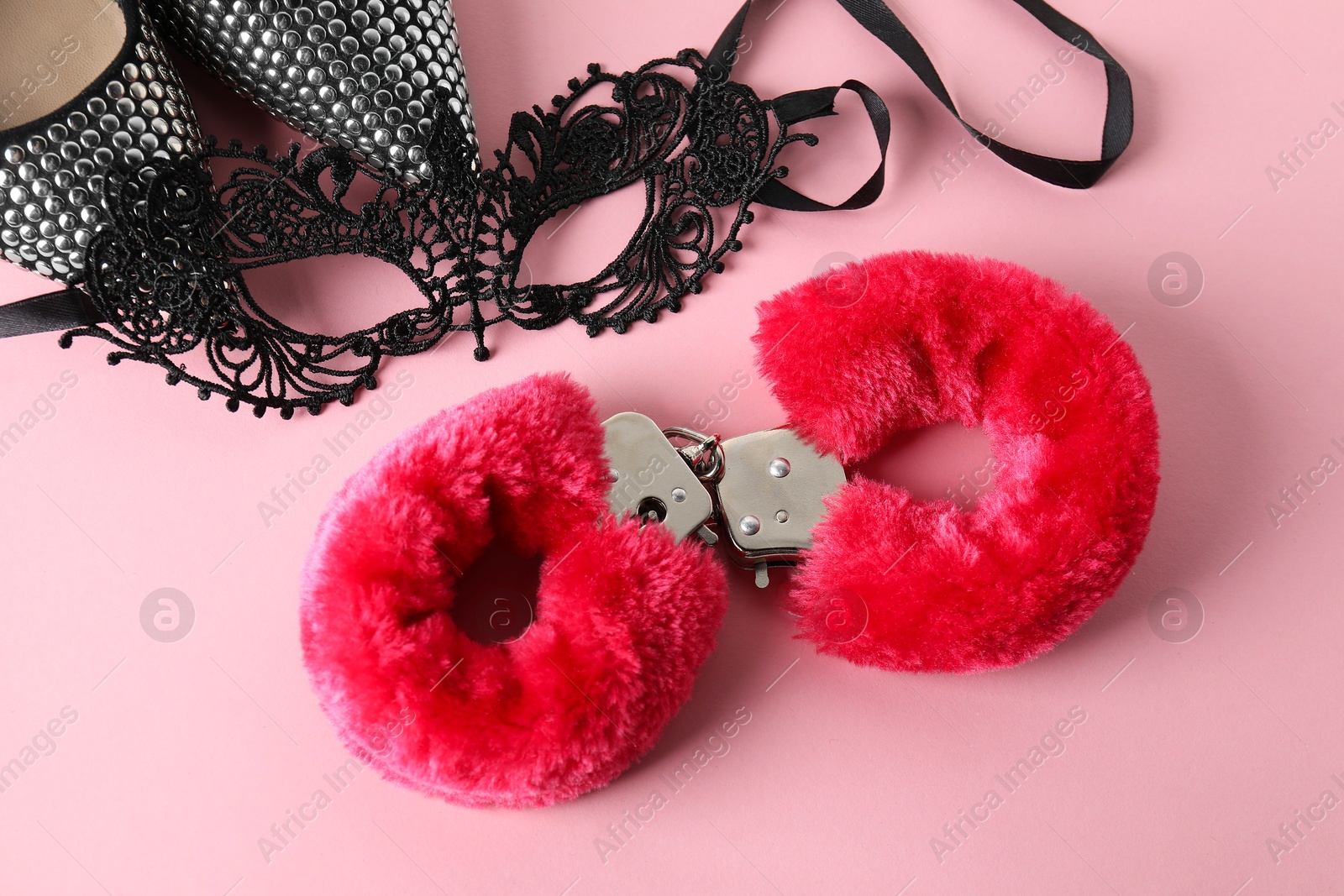 Photo of Fluffy handcuffs, lace mask and high-heeled shoes on pink background, flat lay