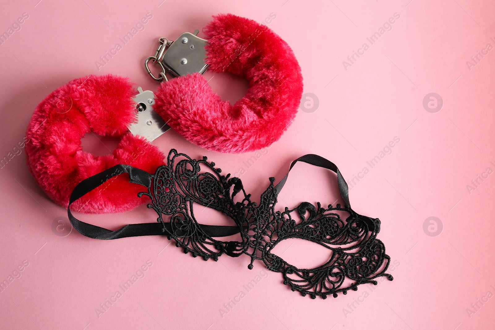 Photo of Fluffy handcuffs and lace mask on pink background, flat lay