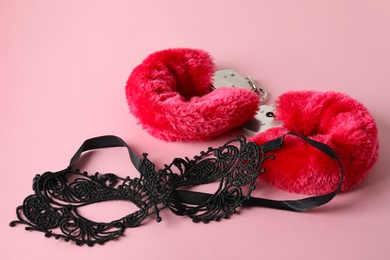 Photo of Fluffy handcuffs and lace mask on pink background, closeup