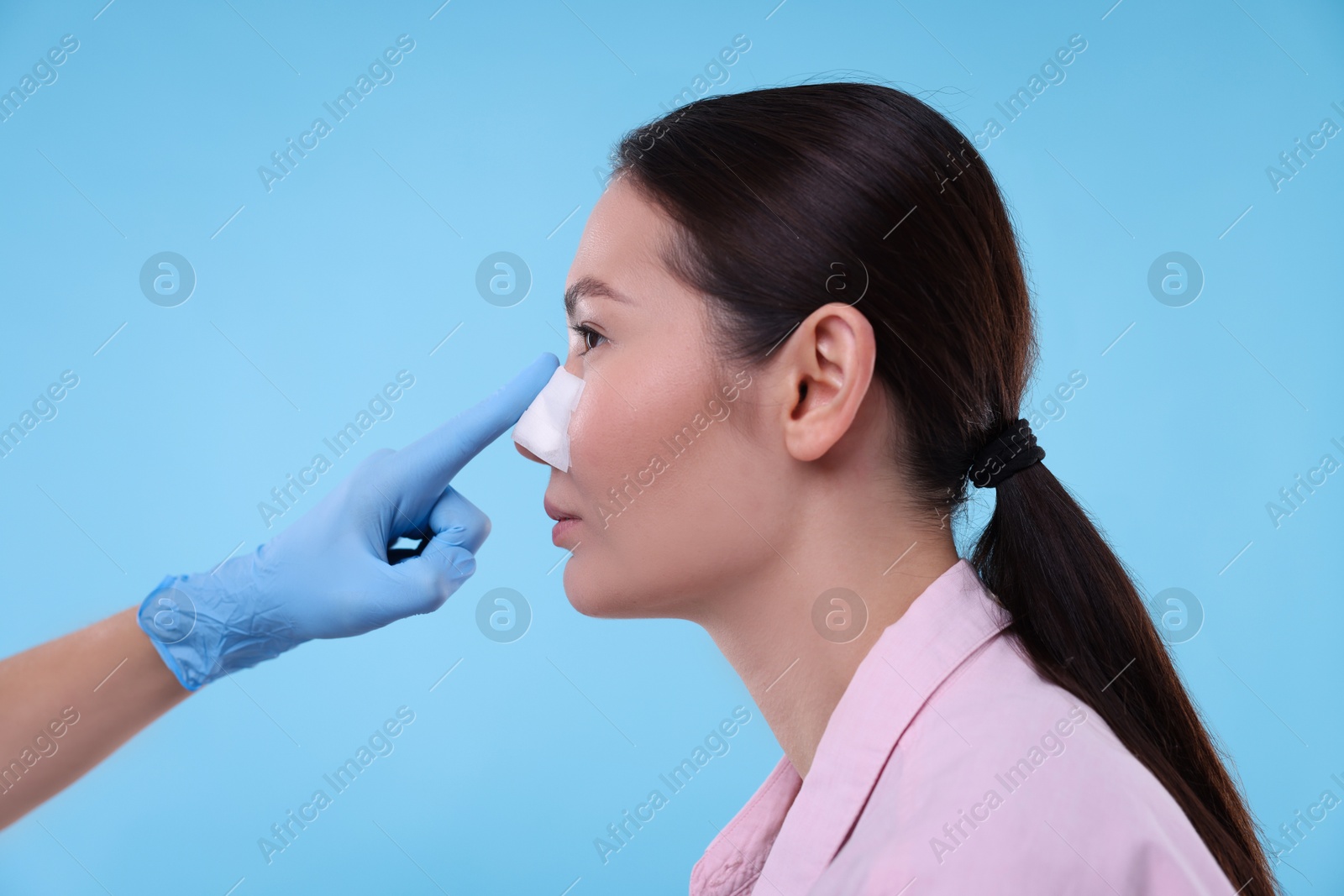 Photo of Doctor checking patient's nose after plastic surgery operation on light blue background, closeup