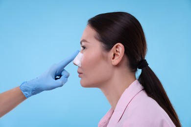 Photo of Doctor checking patient's nose after plastic surgery operation on light blue background, closeup