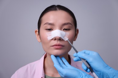 Photo of Doctor removing medical bandage from patient's nose after plastic surgery operation on light grey background, closeup