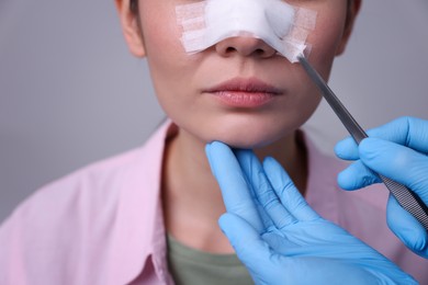 Photo of Doctor removing medical bandage from patient's nose after plastic surgery operation on light grey background, closeup