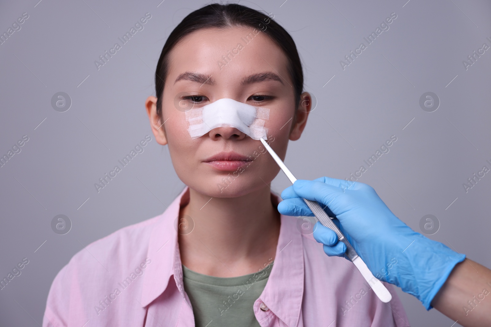 Photo of Doctor removing medical bandage from patient's nose after plastic surgery operation on light grey background, closeup