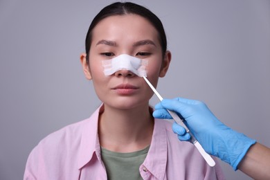 Photo of Doctor removing medical bandage from patient's nose after plastic surgery operation on light grey background, closeup