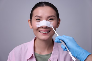 Photo of Doctor removing medical bandage from patient's nose after plastic surgery operation on light grey background, closeup