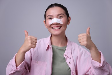 Photo of Woman with medical bandage on her nose after plastic surgery operation showing thumbs up against light grey background