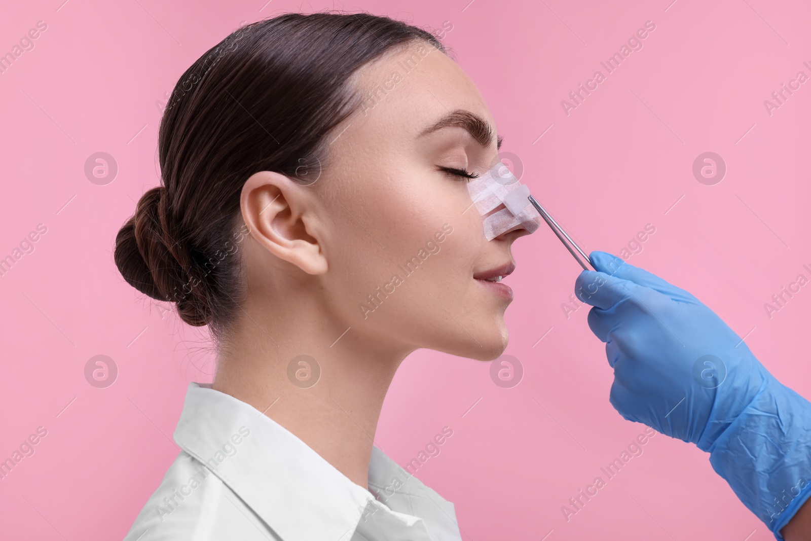 Photo of Doctor removing medical bandage from patient's nose after plastic surgery operation on pink background, closeup