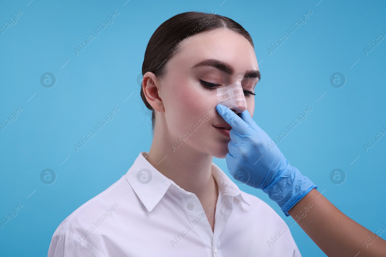 Photo of Doctor checking patient's nose after plastic surgery operation on light blue background, closeup